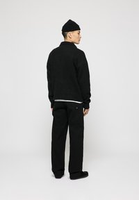Schwarzer Strickpullover mit hohem Kragen, kombiniert mit schwarzen Cargo-Hosen mit Seitentaschen. Das Model trägt eine schwarze Mütze und flache Schuhe.
