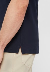 Męskie ramię obok granatowego, krótkiego rękawa koszulki z bocznym rozcięciem, widoczna kieszeń beżowych spodni na jednolitym tle.