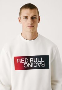 Biele mikina so zaobleným výstrihom a jemnou textúrou, ktorá obsahuje obdĺžnikovú grafiku s červenou až čiernou gradientnou farbou a textom "RED BULL RACING" v tučných bielych písmenách.