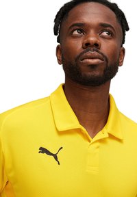 Geel polo shirt van textuurstof met een zwart Puma-logo op de linkerborst. Klassieke kraag en drie knopen.