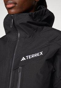 Schwarze Terrex-Jacke mit hohem Kragen, Frontreißverschluss und weißer Logo-Stickerei auf der Brust. Glattes Material mit matter Oberfläche.