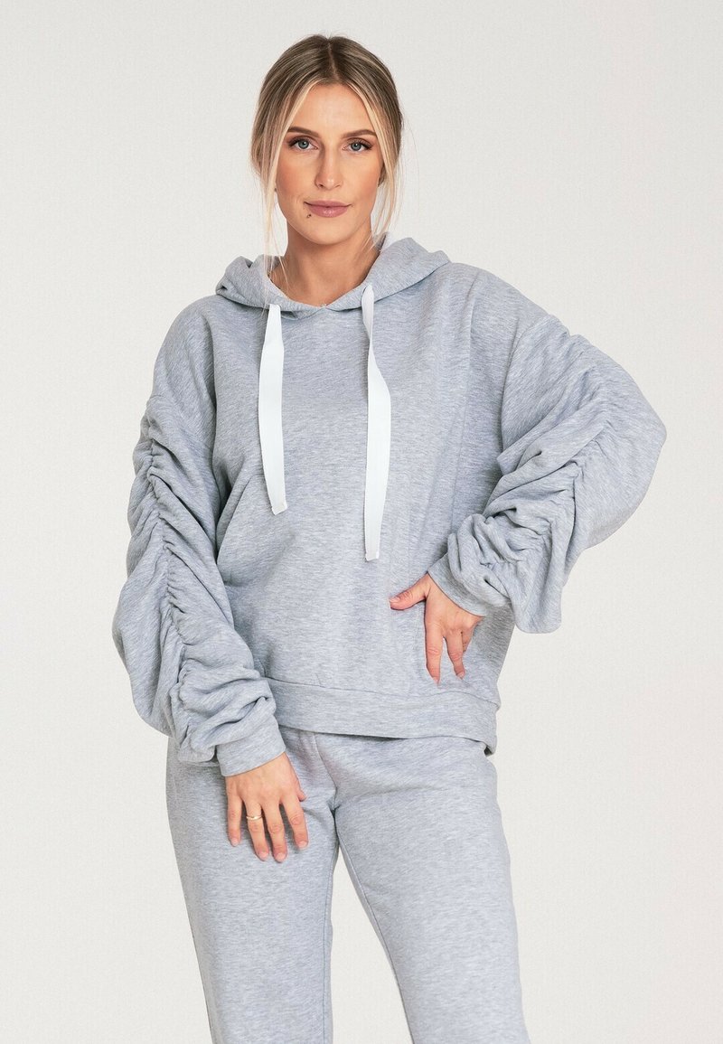 Szara bluza z kapturem z białymi sznurkami i marszczonymi rękawami. Miękka tkanina, luźny krój, w zestawie z dopasowanymi szarymi spodniami dresowymi.