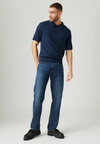 Marineblaues Polo mit kurzen Ärmeln, Kragen und drei Knöpfen, kombiniert mit Jeans in mittlerem Wascheffekt und schwarzen Schuhen. Glattes Material und entspannte Passform.