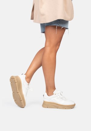 Nogi w dżinsowych szortach i beżowej kurtce, w białych trampkach z grubymi beżowymi podeszwami, jedna stopa lekko uniesiona, ukazująca spód buta.