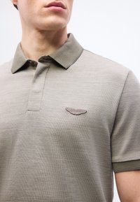 Άνδρας που φοράει ανοιχτό πράσινο-ελιάς πόλο μπλουζάκι με υφασμάτινη υφή και λογότυπο φτερών Aston Martin στο αριστερό στήθος.