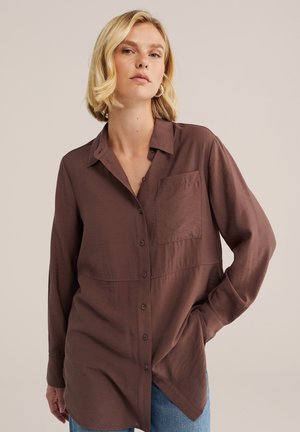 Blond kvinde iført en brun langærmet skjorte med brystlomme og blå jeans, står med den ene hånd i lommen.