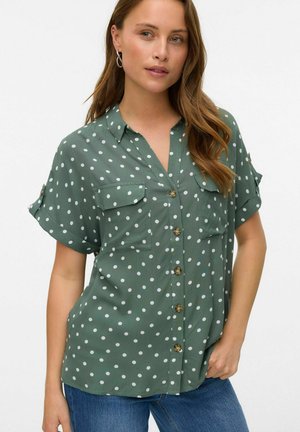 Blusa verde a pois con chiusura frontale a bottoni, due tasche sul petto e maniche arrotolate. Tessuto morbido con una vestibilità rilassata.