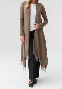 Largo cardigan de punto beige con frente abierto, tejido texturizado y dobladillo con flecos, que se lleva sobre una blusa blanca y pantalones negros.