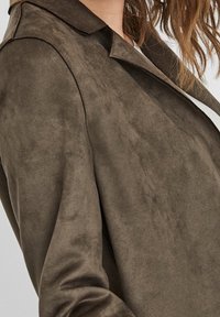 Ruskea mokkajakku, jossa on pehmeä rakenne, perinteinen kaulus ja mukautettu istuvuus. Näkyvät ompeleet korostavat muotoilua.