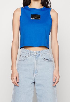Kobieta ubrana w bezrękawnikowy królewsko niebieski krótki top z logo "Karl Lagerfeld Jeans" oraz jasnoniebieskie, szerokie spodnie z wysokim stanem.