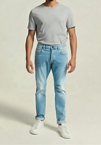 Homme portant un t-shirt gris clair uni, un jean bleu clair avec revers et des baskets blanches, debout devant un fond uni.