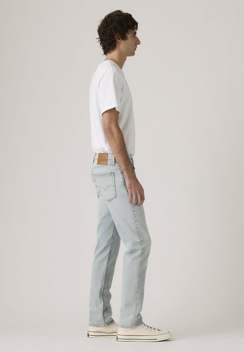 Man in white t-shirt, light blue jeans, and white sneakers standing in profile against a plain background.