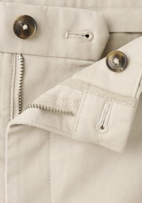 Tessuto di cotone beige con cerniera, due bottoni e asole. Le cuciture sono visibili lungo i bordi del tessuto.