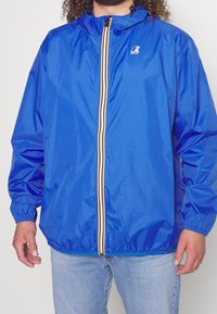 Blauwe waterdichte jas met ritssluiting aan de voorkant en een wit en geel streepje. Elastische manchetten en een capuchon met verstelbare koordjes.