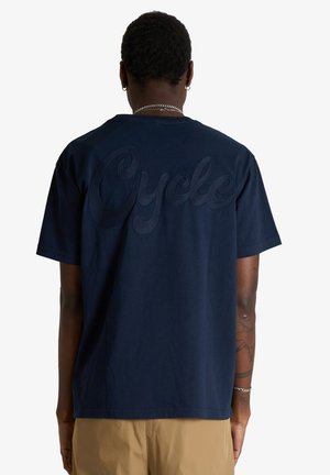 Uomo di spalle che indossa una maglietta blu navy con scritta strutturata "Cycle" sulla schiena e pantaloni beige, mostrando tatuaggi sulle braccia e gioielli in argento.