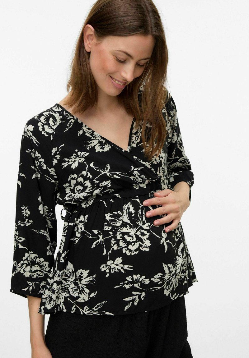 Top portefeuille floral noir avec des manches 3/4, présentant des motifs de fleurs blanches. Il se resserre à la taille, offrant un ajustement confortable. Texture de tissu douce.