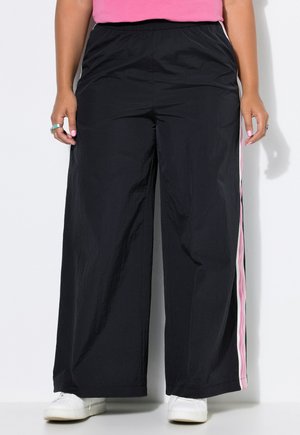 Personne portant un pantalon noir large avec des bandes latérales roses et blanches, des baskets blanches et un haut rose sur fond blanc.