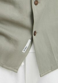 Camisa de llevar verde oliva con una textura suave, que cuenta con botones marrones y una etiqueta de tela blanca. Diseñada con un ajuste entallado.