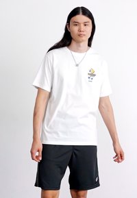 Hvid bomulds-t-shirt med et farverigt grafisk design på venstre bryst, parret med sorte atletiske shorts.