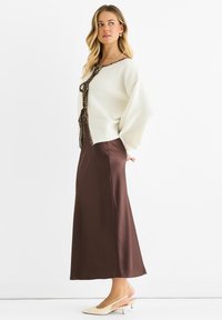 Maglione crema con cravatta a stampa leopardata, abbinato a una lunga gonna di raso marrone e décolleté crema con tacco. Texture morbide e vestibilità rilassata.