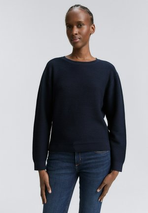 Pull en maille bleu marine avec un col rond et des manches longues, doté de poignets et d'un ourlet côtelés, porté sur un jean bleu.