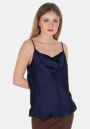 Giovane donna con capelli lisci castano chiaro, che indossa un top senza maniche blu navy con spalline sottili e una gonna marrone, posa contro uno sfondo bianco.