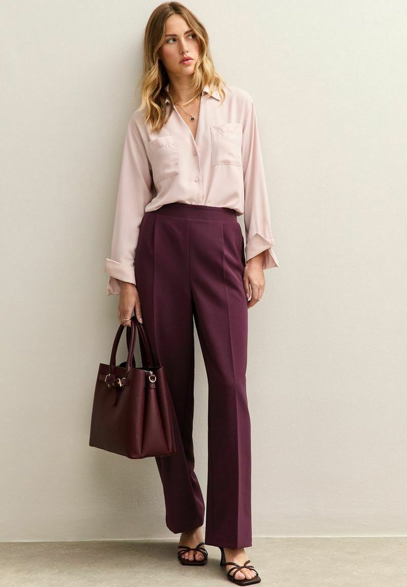 Lichtroze blouse met knopen en twee voorzakken, gecombineerd met hoog-gewijze burgundy broek met wijde pijpen. Inclusief een donker burgundy handtas.
