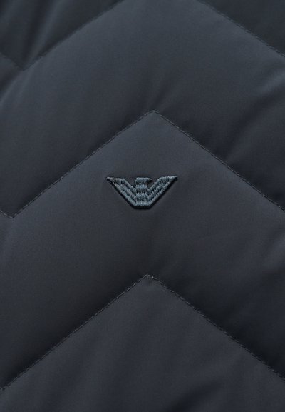 Σκούρο υφασμάτινο καπιτονέ με ραφές σχήματος ζιγκ-ζαγκ και ένα μικρό κεντητό έμβλημα στο κέντρο που μοιάζει με στυλιζαρισμένα φτερά.