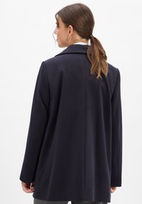 Donkerblauwe jas met een op maat gemaakt ontwerp, met geknipte revers en lange mouwen. De gladde textuur en de relaxte pasvorm complementeren de minimalistische stijl.