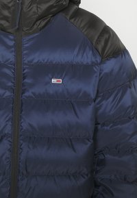 Mörkblå puffjacka med svarta axlar, med en framdragkedja och en liten logopatch på bröstet. Slät, glänsande yta.