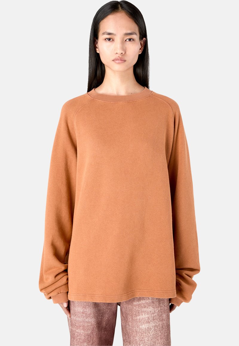 Oversized orange-brown sweatshirt with a rounded neckline, long sleeves, ribbed cuffs, and soft, textured fabric. No visible logos or patterns.