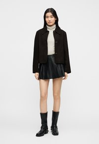 Γυναίκα που φοράει μαύρο σακάκι με κουμπιά, λευκή ζιβάγκο με ρίγες, μαύρη πλισέ μίνι φούστα και μαύρες μπότες μέχρι τη μέση της γάμπας, στέκεται μπροστά σε απλό φόντο.