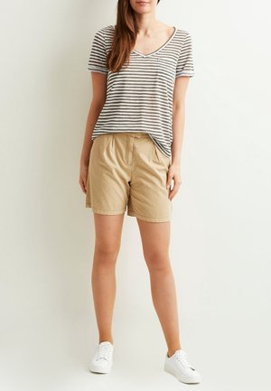 Femme portant un t-shirt à manches courtes rayé gris et blanc, un short beige et des baskets blanches, debout devant un fond clair uni.