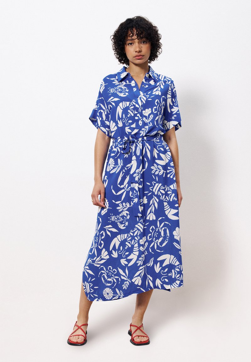 Blaues Midikleid mit weißem Blumenmuster, kurzen Ärmeln, Knopfleiste und Taillengürtel. Kombiniert mit roten Riemchensandalen.