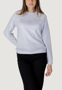 Sudadera de color púrpura claro con cuello redondo, puños acanalados y un diseño de logotipo sutil en la parte delantera. Combinada con pantalones negros.