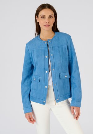 Femme portant une veste en daim bleue à boutons-pression et fermeture éclair, associée à un pantalon blanc, sur un fond uni clair.