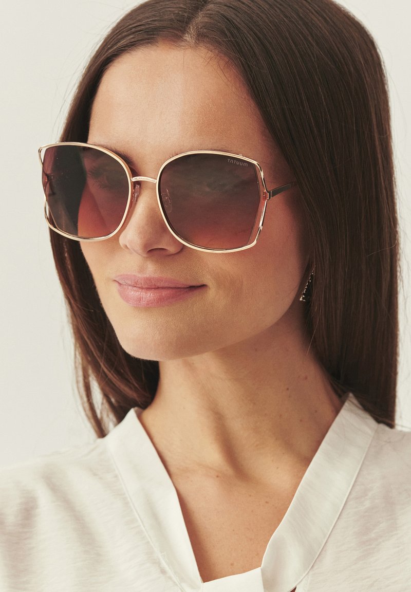 Femme aux cheveux longs et raides bruns, portant de grandes lunettes de soleil carrées marron dégradé et un haut blanc sans col, tournée légèrement vers la droite.