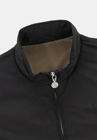 Svart jacka med hög krage, kontrasterande tanfärgad insida, frontdragkedja med metalldragflik och en cirkulär logotyp på vänster sida.