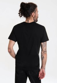 Black cotton t-shirt with short sleeves, round neckline, and a smooth texture. Visible tattoos on arms, worn with black jeans.