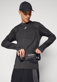Black long-sleeve athletic top with textured fabric, featuring a logo on the chest. The wearer adjusts a black running belt with flasks.