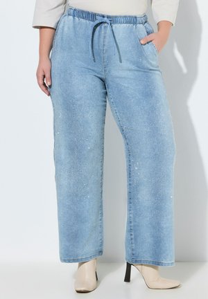 Kvinde iført lyseblå viddebukser med glitrende detaljer, elastisk talje med snøre, cremefarvede ankelstøvler med hæl og en hvid top.