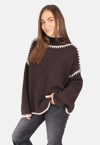 Dunkelbrauner Strickpullover mit hohem Kragen, weißem gewellten Saum, weiter Passform und weiten Ärmeln, kombiniert mit grauer Jeans.