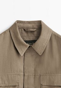 Gros plan sur le col d'une chemise en lin beige avec patte de boutonnage et deux poches poitrine à rabats sur fond blanc.