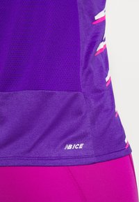 Fialový sportovní top s detaily z síťoviny, hladkou texturou a bílými a růžovými proužky. Na lemu je logo.