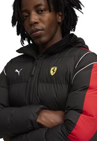 Zwarte gewatteerde jas met rode mouwaccenten, Puma-logo en Ferrari-embleem. Beschikt over een staande kraag en een ritssluiting aan de voorkant. Gladde textuur.
