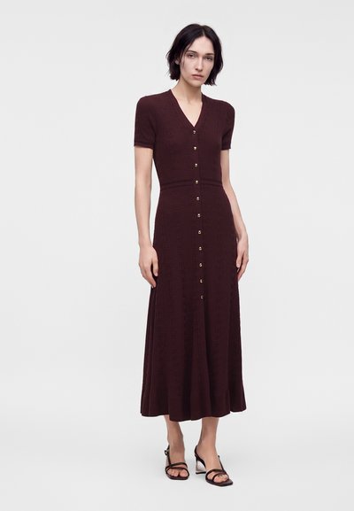 Femme portant une robe en maille bordeaux foncé à boutons, manches courtes, et des talons ouverts noirs, debout devant un fond clair uni.