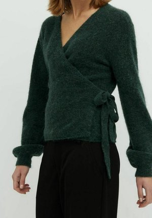 Kvinde iført en mørkegrøn, blød wrap-trøje med lange ærmer bundet ved taljen, kombineret med sorte bukser, stående mod en ensfarvet baggrund.