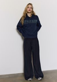 Mornarskoplava majica s tekstom "STYLE ICON" od kristala, u kombinaciji sa širokim crnim hlačama i srebrnim tenisicama, na neutralnoj pozadini.