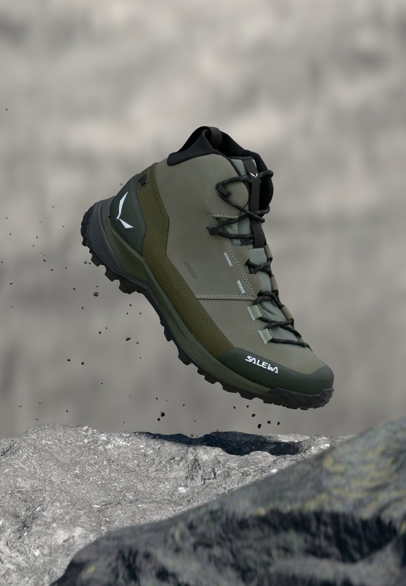 Botte de randonnée imperméable verte et noire avec lacets, modèle mi-haut, montrée en plein saut au-dessus d'un terrain rocheux avec des particules de terre tombant.