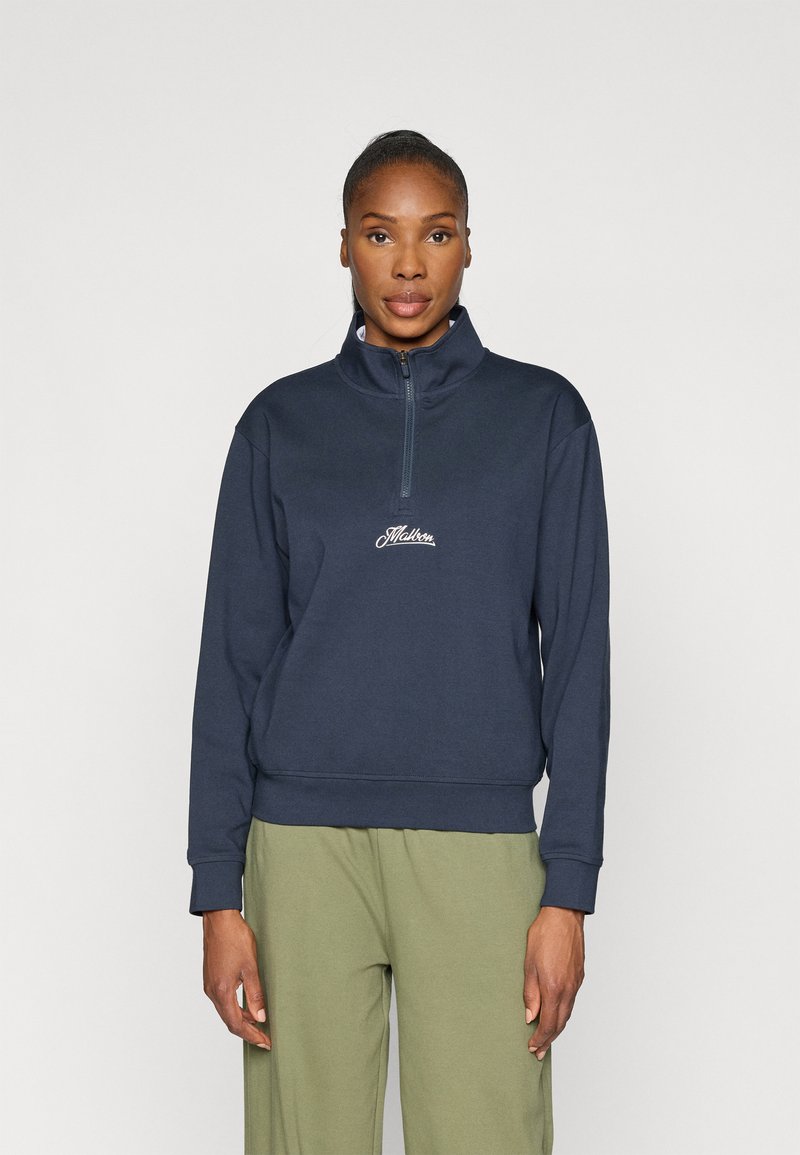 Zilā kapučsweatshirt ar rāvējslēdzēju, augstu apkakli un izšūtu logotipu uz krūtīm, ar ribotām piedurknēm un apakšmalu, kombinēts ar olīvu zaļām biksēm.